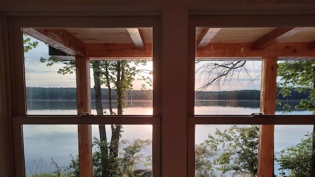 Ein Zimmer mit großen Fenstern, die auf einen See blicken, Holzbalken und Blick auf Bäume und Hügel.