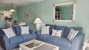 Ein Wohnzimmer mit zwei blauen Sofas, einem Glastisch, einer weißen Lampe und einem Spiegel an der Wand.