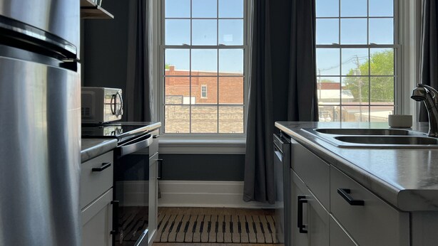 Eine moderne Küche mit einem Edelstahlkühlschrank, einer Mikrowelle und einem Spülbecken. Zwei Fenster mit Vorhängen, ein Teppich auf dem Boden und eine hängende Leuchte sind vorhanden.