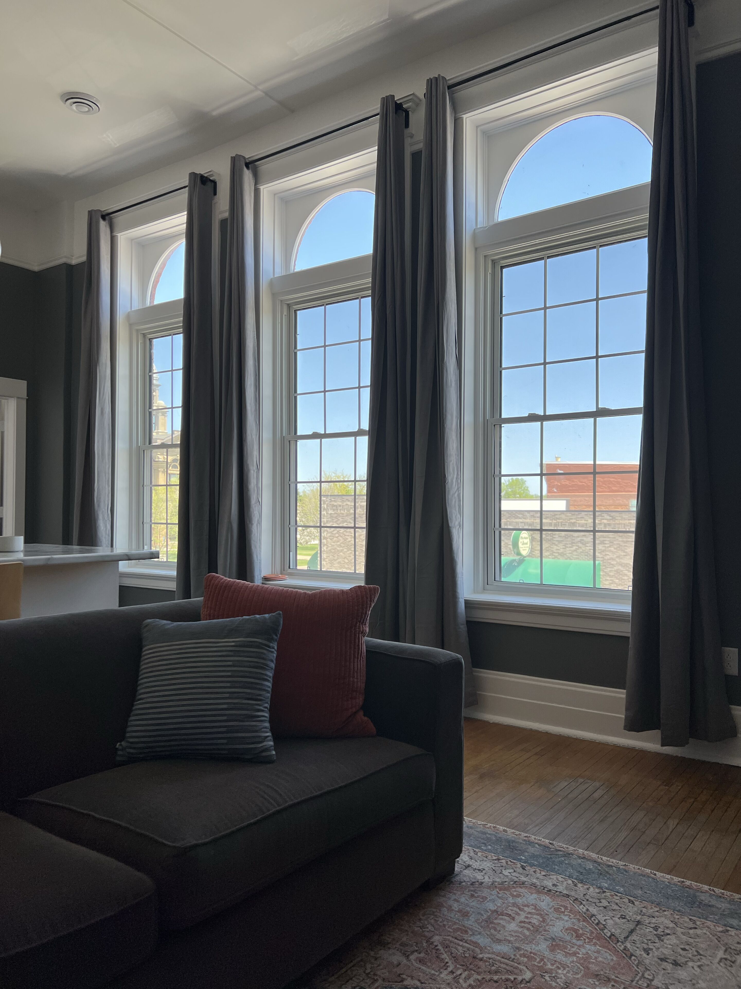 A living room with a dark-colored sofa, a patterned rug, and large windows with curtains.