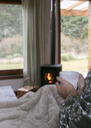 A person holding a cup, sitting by a window with a view of greenery, and a wood-burning stove in the background. - Vale Mandaçaia (Urubici)