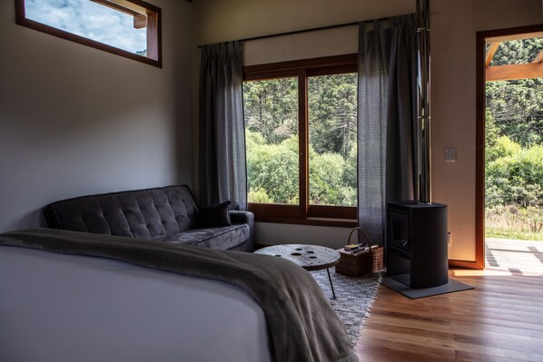 Un dormitorio con una cama grande, un sofá, una pequeña mesa redonda, una chimenea y vistas al verdor por la ventana.