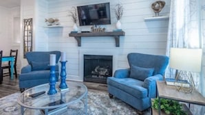 Una sala de estar con dos sofás azules, una mesa de centro de cristal, una chimenea y un televisor montado.