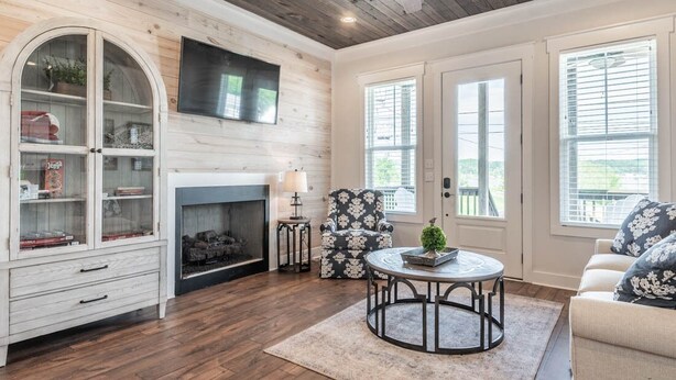 A living room with a fireplace, a television mounted on the wall, a ceiling fan, and a large window.