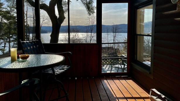 Una cabaña acogedora con una terraza de madera, una mesa redonda con sillas y un amplio ventanal que ofrece vistas a un lago y árboles.