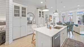 A modern kitchen with white cabinetry, a central island, pendant lighting, and a dining area with wicker chairs. - Designer Cinnamon Shore Condo w/ Charm+Easy Access to Countless Amenities! (Port Aransas)