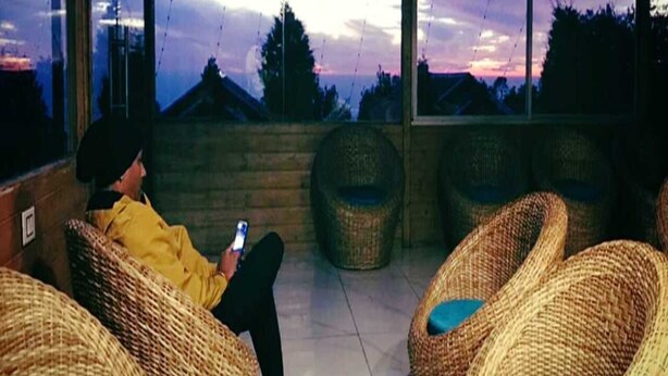 A person sitting on a wicker chair with a smartphone, overlooking a sunset through large windows. - Katyuri Nature View Resort (Lansdowne)