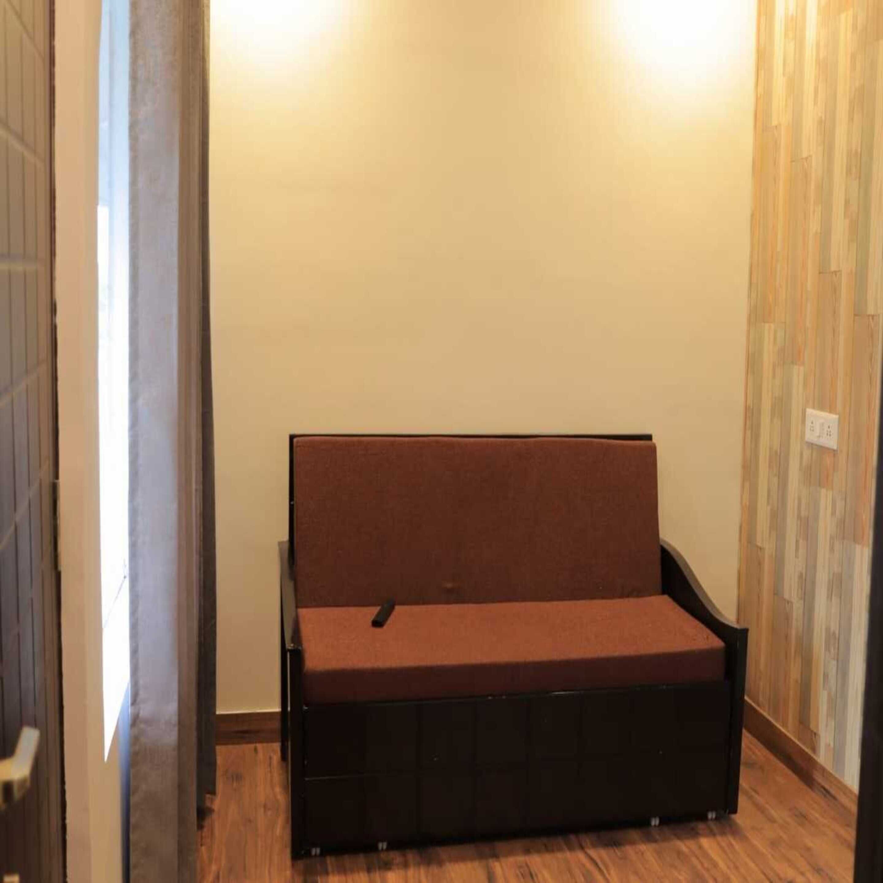 A small, dark-colored sofa in a narrow hallway with wooden flooring and a plain wall.