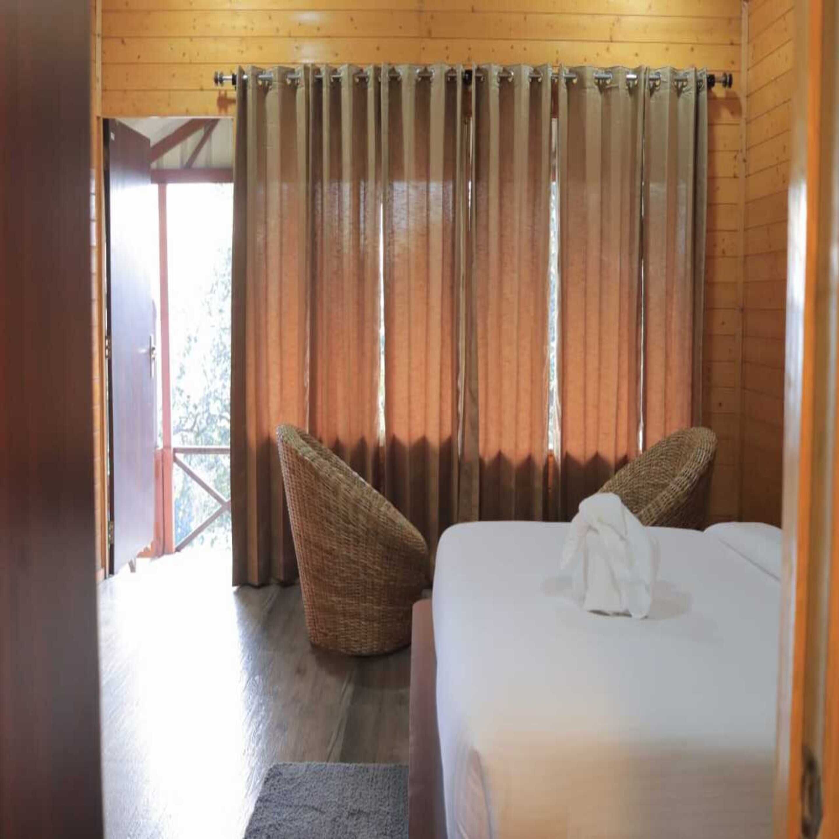 A bedroom with a bed, two wicker chairs, wooden walls, and a view through a door.
