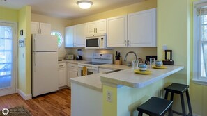 A kitchen with white cabinets, a microwave, a stove, and a sink. There is a window with blinds and a door on the left.