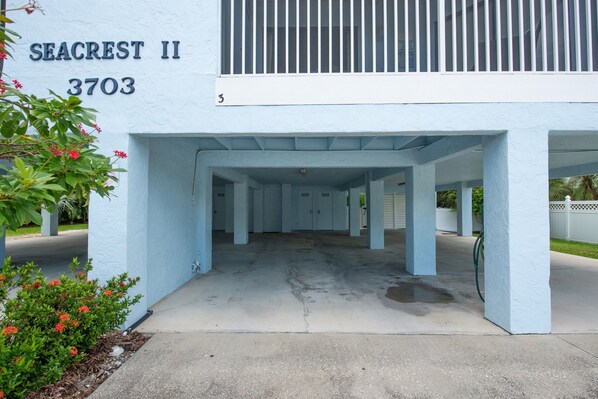 A covered parking area with a light blue building, a number '3703' on the wall, and some greenery.