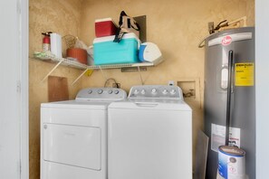 A laundry room with two white front-loading washers, a shelf with various items, and a large water heater.