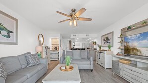 A modern living room with a ceiling fan, a grey sofa, a white coffee table, and a kitchen in the background. - Beach House - Sky’s the Limit (Panama City Beach)