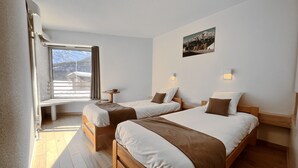 A hotel room with two beds, a window with a view of mountains, a small table, and a wall-mounted picture. - La Bessannaise (Bessans)