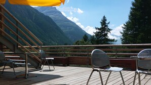 A wooden deck with metal chairs and a yellow umbrella, overlooking a mountainous landscape. - La Bessannaise (Bessans)
