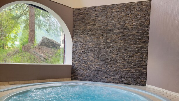 A spa area with a hot tub, a stone wall, and a view of greenery through a large window. - La Bessannaise (Bessans)