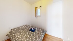 Una habitación pequeña y escasamente amoblada, con una colcha estampada, una ventana con cortina y piso de madera.