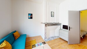 Una habitación pequeña con un sofá azul, una mesita con una revista, un televisor en un mueble y una puerta que da a otra habitación.