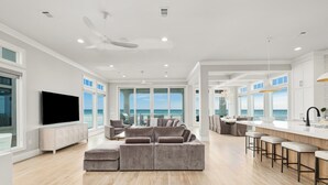 A spacious living area with a large sectional sofa, a flat-screen TV, and a view of the ocean through large windows. - Gulf-Front with Rooftop Deck, Theater and Pool (Miramar Beach)