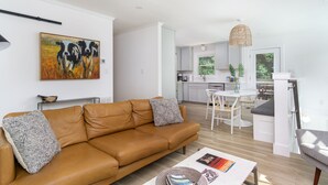A modern apartment with a living room featuring a brown leather sofa, a dining area with a white table and chairs, and a kitchen with light grey cabinets. - Cottontail Cottage | Pet-friendly, Fire Pit & 10 Min. to Downtown AVL! (Asheville)