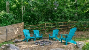 A garden with four turquoise Adirondack chairs arranged around a small fire pit, surrounded by rocks and a wooden fence. - Cottontail Cottage | Pet-friendly, Fire Pit & 10 Min. to Downtown AVL! (Asheville)