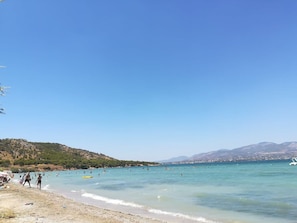 Uma praia com águas azul-claras, pessoas nadando e uma paisagem montanhosa ao fundo.