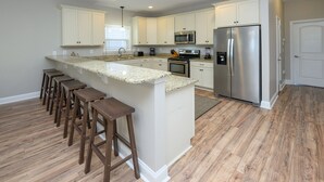 Eine moderne Küche mit weißen Schränken, einem Edelstahl-Kühlschrank, einer Mikrowelle und einer Arbeitsplatte aus Granit. An der Kücheninsel stehen Holzbarhocker.