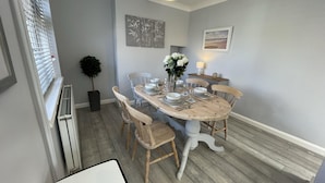 Un coin repas avec une table ronde en bois, dressée pour quatre personnes, entourée de chaises en bois. Un pot de plante se trouve à gauche et une petite table avec une lampe, à droite.