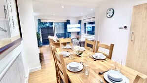 Un coin repas avec une table en bois dressée pour quatre personnes, des chaises, une horloge au mur et une vue sur un jardin par la fenêtre.