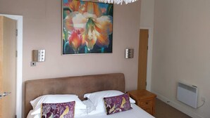 Une chambre à coucher avec un lit, deux oreillers, une table de chevet, un luminaire fixé au mur et un tableau représentant des fleurs.