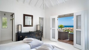 Een slaapkamer met een bed, een kaptafel en een grote spiegel. Een deur leidt naar een balkon met uitzicht op zee en gebouwen.