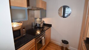 Eine kompakte Küche mit Mikrowelle, Toaster und Wasserkocher auf der Arbeitsfläche, Holzschränken und einer kleinen Mülltonne.