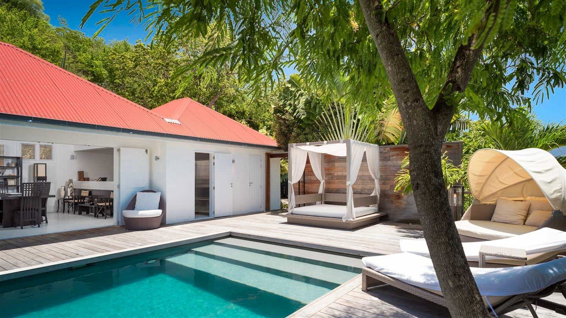 Un espace aménagé au bord de la piscine, avec une terrasse en bois, un bâtiment au toit rouge, un coin salon couvert et un lit à baldaquin blanc.