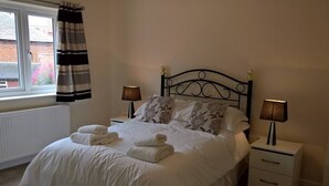 Un lit bien fait, avec du linge de lit blanc et des serviettes pliées, deux lampes de chevet, une fenêtre avec des rideaux et un radiateur.