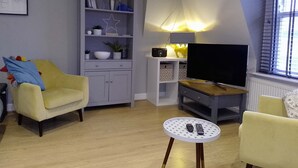 Ein Wohnzimmer mit einem gelben Sessel, einer grauen Schrank mit Regalen, einem Fernseher auf einem Holzständer und einem kleinen runden Tisch mit einer Fernbedienung.
