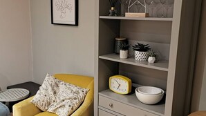 Ein gelber Sessel mit einem gemusterten Kissen, eine graue Regaleinrichtung mit Dekoration und Pflanzen sowie ein kleiner runder Tisch mit weißer Tischdecke.