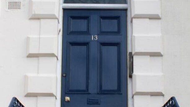 Eine dunkelblaue Tür mit der Nummer 13, begleitet von zwei Blumentöpfen, die zu einer Treppe mit weißer Brüstung führt.