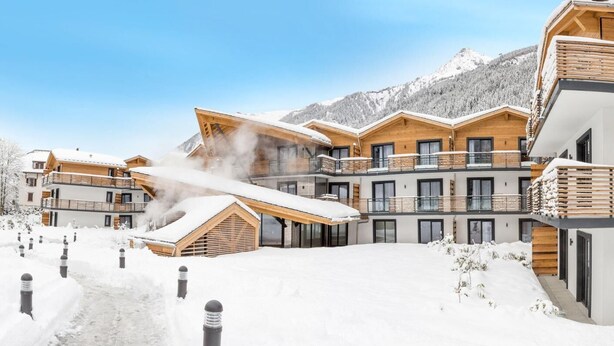A modern ski resort with wooden buildings, snow-covered balconies, and a clear view of snow-covered mountains in the background. - Wifi, fitness, swimming pool, sauna, hammam, parking, tv, 34m², Chamonix (Chamonix-Mont-Blanc)