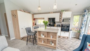 A modern kitchen with a central island, built-in appliances, and a dining area with a grey sofa and bar stools. - Loluke Lodge on The Lake (Kendal)