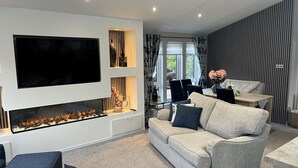 A modern living room with a flat-screen TV, a fireplace, a sofa, and a dining area. - Loluke Lodge on The Lake (Kendal)