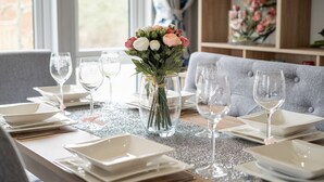 A dining table set with white plates, cutlery, and wine glasses, with a vase of flowers in the center. - Loluke Lodge on The Lake (Kendal)