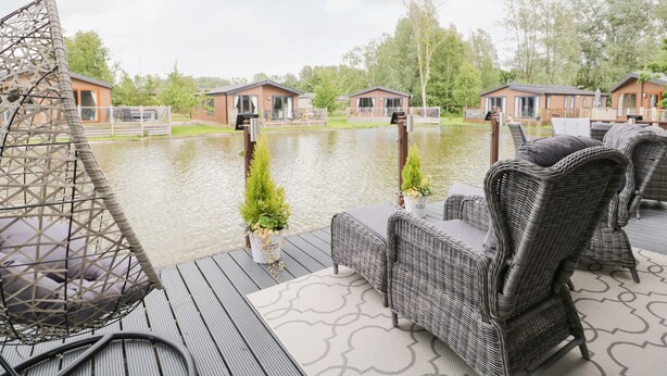 A deck with wicker furniture and a swing chair overlooking a body of water with houses in the background. - Loluke Lodge on The Lake (Kendal)