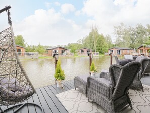 En terrass med möbler av rotting och en hängstol med utsikt över en vattenyta, med hus i bakgrunden.