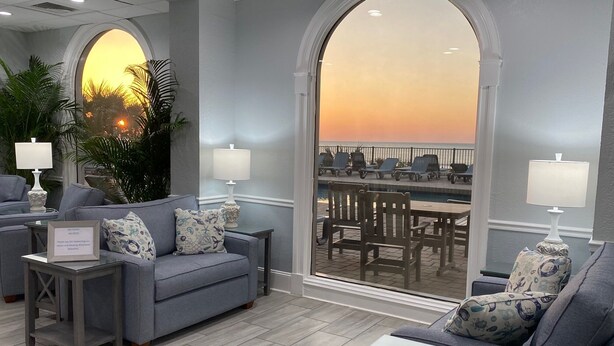 Uma área de estar moderna com sofá, poltronas e mesa de vidro. Há duas luminárias, uma placa e vista para a área da piscina externa.