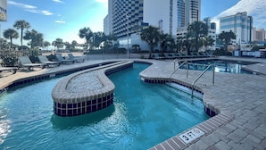 Piscina com indicador de profundidade de 3 pés, rodeada por cadeiras de praia e palmeiras, com edifícios modernos ao fundo.