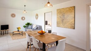 Un comedor moderno con una mesa de madera, sillas blancas y un frutero. Hay un televisor sobre un mueble bajo, un sofá y un cuadro abstracto grande en la pared.