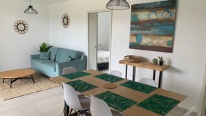 Ein Essbereich mit einem Holztisch, weißen Stühlen und grünen Platzsets. Ein hellblaues Sofa und ein kleiner runder Tisch mit einem gewebten Platzset sind ebenfalls vorhanden. Über dem Esstisch hängt ein Gemälde.