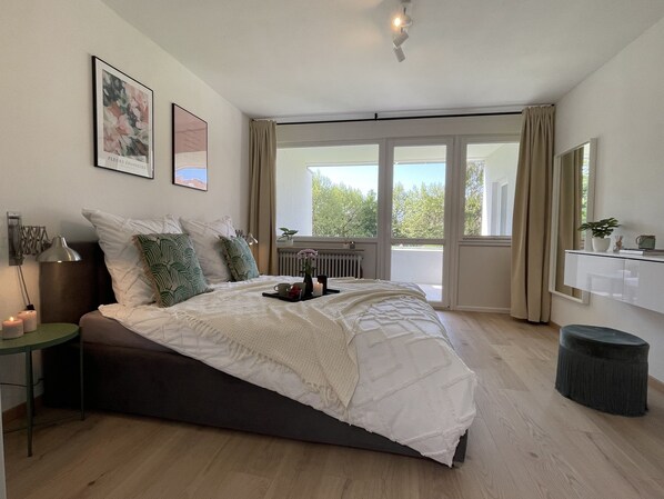 A bedroom with a large bed, a bedside table, a nightstand, a window with curtains, and a view of greenery outside.
