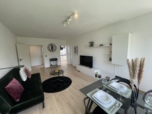 Una sala moderna con un sofá, una mesa de comedor para dos personas, un televisor de pantalla plana y una cocina al fondo.