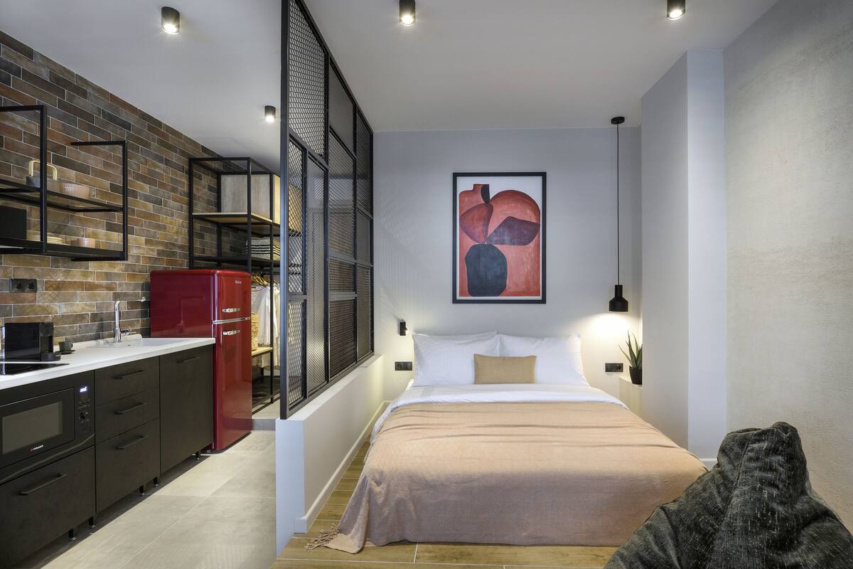 A modern bedroom with a large bed, a red refrigerator, a kitchen area with a brick wall, and a large abstract painting on the wall.